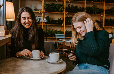 Junge Frauen lachen über Witze, während sie Kaffee trinken und ihr Smartphone während eines Treffens in einem gemütlichen Restaurant benutzen - ADSF11442