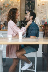 Seitenansicht eines fröhlichen Paares, das sich in einem Café umarmt, ein heißes Getränk trinkt und sich gegenseitig anschaut - ADSF11451