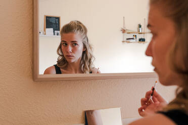 Young female reflected in mirror contouring eyebrows with black cosmetic pencil while doing makeup at home - ADSF11455