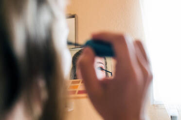 Young female holding palette with colorful eye shadows and mirror and applying black mascara on eyelashes during beauty procedure at home - ADSF11456