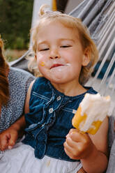 Zufriedenes kleines Kind, das ein selbstgemachtes Eis am Stiel isst, während es sich im Sommer auf der Terrasse entspannt - ADSF11462