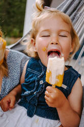 Zufriedenes kleines Kind, das ein selbstgemachtes Eis am Stiel isst, während es sich im Sommer auf der Terrasse entspannt - ADSF11463