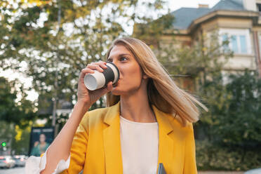 From below trendy slim blond businesswoman looking away in elegant vivid yellow jacket and jeans with digital tablet walking alone against exteriors of residential multistory buildings and cars parked on street in downtown - ADSF11467