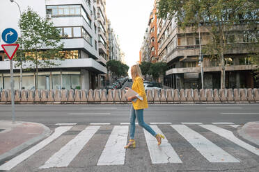 Side view of trendy slim blond female in elegant vivid yellow jacket and jeans with digital tablet walking alone along pedestrian crossing against exteriors of residential multistory buildings and cars parked on street in downtown - ADSF11469