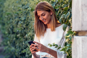 Positive entspannte blonde Frau in Freizeitkleidung, die sich auf den Bildschirm konzentriert und lächelt, während sie auf der Straße steht und mit ihrem Smartphone in der Nähe einer mit einem Zaun bedeckten grünen Kletterpflanze interagiert - ADSF11470