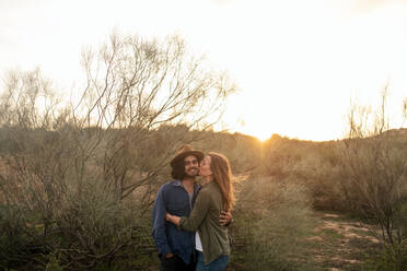 Fröhlicher Mann und Frau, die sich umarmen und sich gegenseitig ansehen, während sie in der Nähe eines blattlosen Busches bei einem abendlichen Date in der Natur stehen - ADSF11474