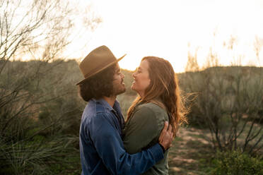 Fröhlicher Mann und Frau, die sich umarmen und sich gegenseitig ansehen, während sie in der Nähe eines blattlosen Busches bei einem abendlichen Date in der Natur stehen - ADSF11476