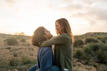 Adult man lifting and embracing optimistic girlfriend while having fun against sundown sky during date in nature - ADSF11482