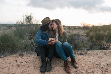 Man in hat sitting on sandy ground and hugging happy girlfriend during romantic date in cloudy evening in countryside - ADSF11488