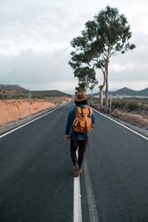 Back view of anonymous man putting on backpack and walking on asphalt road on cloudy day in countryside - ADSF11494