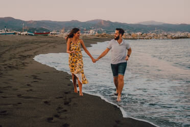 Glückliche junge Leute verlieben sich beim Spazierengehen und halten sich barfuß am Meer im kontrastreichen Sonnenlicht an den Händen - ADSF11498