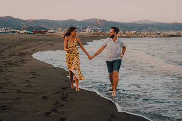 Happy young people fall in love walking looking to each other and holding hands barefoot in seaside in contrast sunlight - ADSF11498