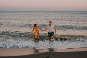Glückliche junge Menschen verlieben sich ineinander, laufen einander zu und halten sich barfuß am Meer im kontrastreichen Sonnenlicht an den Händen - ADSF11500