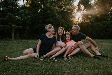 Happy adult couple with adorable daughters sitting on grass near green trees and smiling while resting together and enjoying summer day in nature - ADSF11510