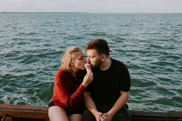 Glückliches junges romantisches Paar in Freizeitkleidung isst Eiswaffel zusammen, während sie auf dem Pier in der Nähe des Meeres sitzen und die Sommerferien zusammen genießen - ADSF11511