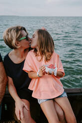 Bezauberndes kleines Mädchen, das eine Schale mit frischen Beeren hält und seine glückliche Mutter küsst, während sie in den Sommerferien zusammen auf einem Zaun am Meer sitzen - ADSF11512