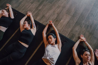 Hoher Winkel einer Gruppe junger sportlicher Frauen und Männer in Sportkleidung mit geschlossenen Augen und gekreuzten Beinen, die sich entspannen und tief atmen, während sie in Savasana-Position liegen und auf Sportmatten in einem modernen Yoga-Studio meditieren - ADSF11517
