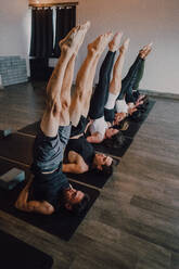 Von oben der Gruppe von erwachsenen sportlichen Menschen in Sportkleidung mit Beinen erhöht Stretching Körper in Salamba Sarvangasana Haltung auf Sportmatten beim Üben von Yoga in modernen Studio - ADSF11518