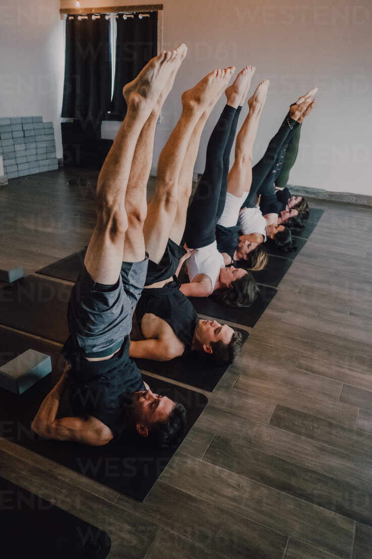 From above of group of adult sporty people in sportswear with legs raised  stretching body in salamba sarvangasana pose on sports mats while  practicing yoga in modern studio stock photo