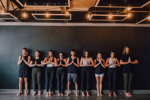 Gruppe von glücklichen jungen sportlichen Menschen in Sportkleidung mit geschlossenen Augen, die sich entspannen und tief atmen, während sie mit Namaste in Tadasana stehen und neben einer schwarzen Wand in einem modernen Yogastudio meditieren - ADSF11521