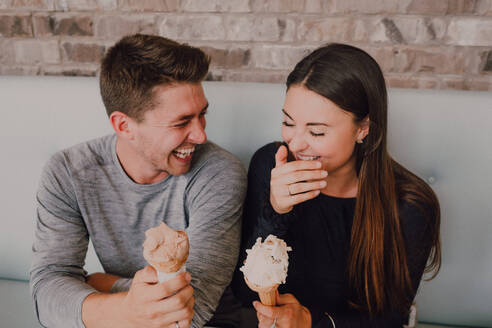 Hohe Winkel von fröhlichen Mann und junge Frau in Freizeitkleidung Blick auf einander mit leckerem Eis, während am Tisch auf dem Sofa sitzen und entspannen zusammen in modernen Café im Loft-Stil - ADSF11527