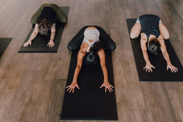 From above unrecognizable ladies in sportswear concentrating and lying in balasana position on sports mats on wooden floor in spacious workout room - ADSF11572