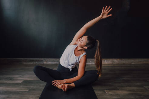 Junge entspannte Frau mit geschlossenen Augen und erhobenem Arm, die in Baddha Konasana Position auf einer Sportmatte sitzt und Yoga in einer dunklen Halle praktiziert - ADSF11576