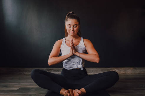 Ruhige, entspannte Frau mit geschlossenen Augen und Namaste, die in Baddha Konasana Position auf einer Sportmatte sitzt und sich konzentriert, während sie in einem dunklen, modernen Studio Yoga übt - ADSF11579
