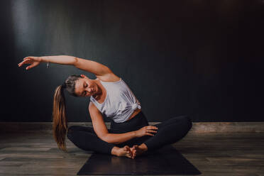 Junge entspannte Frau mit geschlossenen Augen und erhobenem Arm, die in Baddha Konasana Position auf einer Sportmatte sitzt und Yoga in einer dunklen Halle praktiziert - ADSF11580