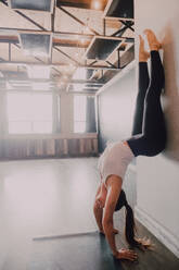 Side view upside down of barefooted female athlete in activewear practicing yoga in downward facing dog pose standing and leaning on white wall of spacious contemporary studio - ADSF11589