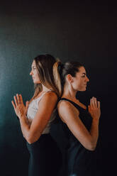 Side view of tranquil relaxed female athletes in activewear with hands folded practicing yoga in tadasana position while standing back to back and looking away against black wall in modern studio - ADSF11592