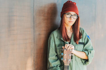 Zufriedene stilvolle Frau mit Brille und dunkelgrünem Hemd, die eine Gitarre hält, steht in der Nähe einer braunen Wand und schaut in die Kamera - ADSF11602