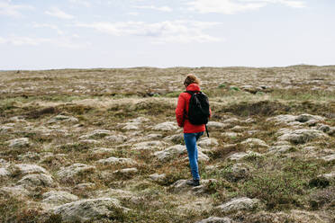 Rückenansicht eines nicht erkennbaren reisenden Mannes in Oberbekleidung und Rucksack, der auf einer felsigen grünen Wiese im Urlaub in Island spazieren geht - ADSF11606