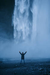 Delighted male tourist in outerwear standing with raised arms on background of waterfall while enjoying vacation in Iceland and looking at camera - ADSF11611