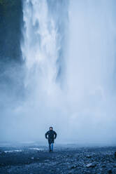 Erfreuter männlicher Tourist in Oberbekleidung, der vor dem Hintergrund eines Wasserfalls spazieren geht, während er seinen Urlaub in Island genießt und in die Kamera schaut - ADSF11612