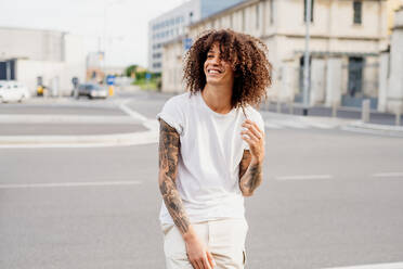 Smiling man with tattooed arms and long brown curly hair standing on a street. - CUF56297