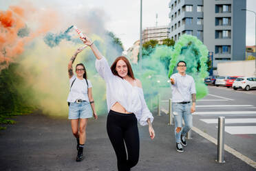 Mixed race group of friends hanging out together in town, using colourful smoke bombs. - CUF56303