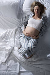 Porträt einer hochschwangeren Frau, die auf dem Bett liegt und den Bauch streichelt. - CUF56317