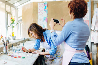 Künstlerin malt auf Stoff im Kreativstudio, reife Frau fotografiert sie mit Smartphone - CUF56341