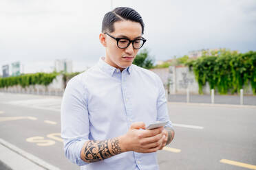 Portrait of man with tattooed arm, wearing light blue shirt and glasses, using mobile phone. - CUF56410