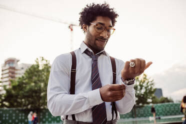 Portrait of businessman wearing glasses, white shirt, brown tie and suspenders, adjusting his cuffs. - CUF56454