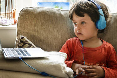 Young boy with brown hair wearing blue headphones, watching the screen of a laptop. - CUF56455