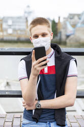 Junger Mann mit Gesichtsmaske bei der Nutzung eines Smartphones in einer Stadt während des Ausbruchs des Coronavirus - WPEF03280