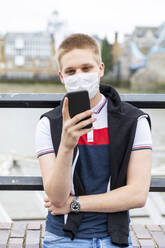 Young man wearing face mask while using smart phone in city during coronavirus outbreak - WPEF03280