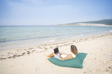 Mann benutzt Laptop und entspannt sich auf einem großen Kissen am Strand, Tarifa, Spanien - OCMF01648