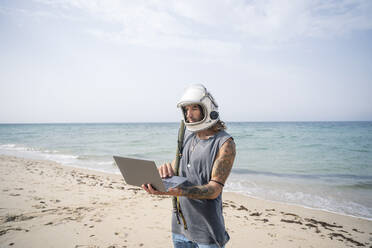 Man in space helmet using laptop while standing at beach, Tarifa, Spain - OCMF01649