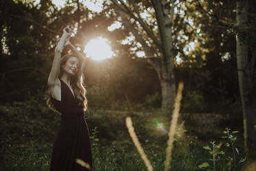 Smiling woman with arms raised against sunlight standing in forest - GMLF00449