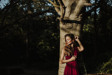 Lächelnde Frau mit geschlossenen Augen, die an einem Baumstamm im Wald steht - GMLF00453