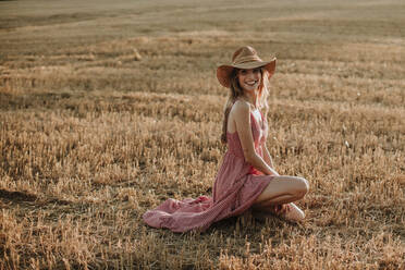 Lächelnde junge Frau kniend in einem Weizenfeld bei Sonnenuntergang - GMLF00455