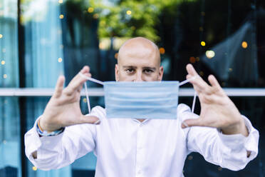 Bald man holding protective face mask while standing outdoors - DGOF01238